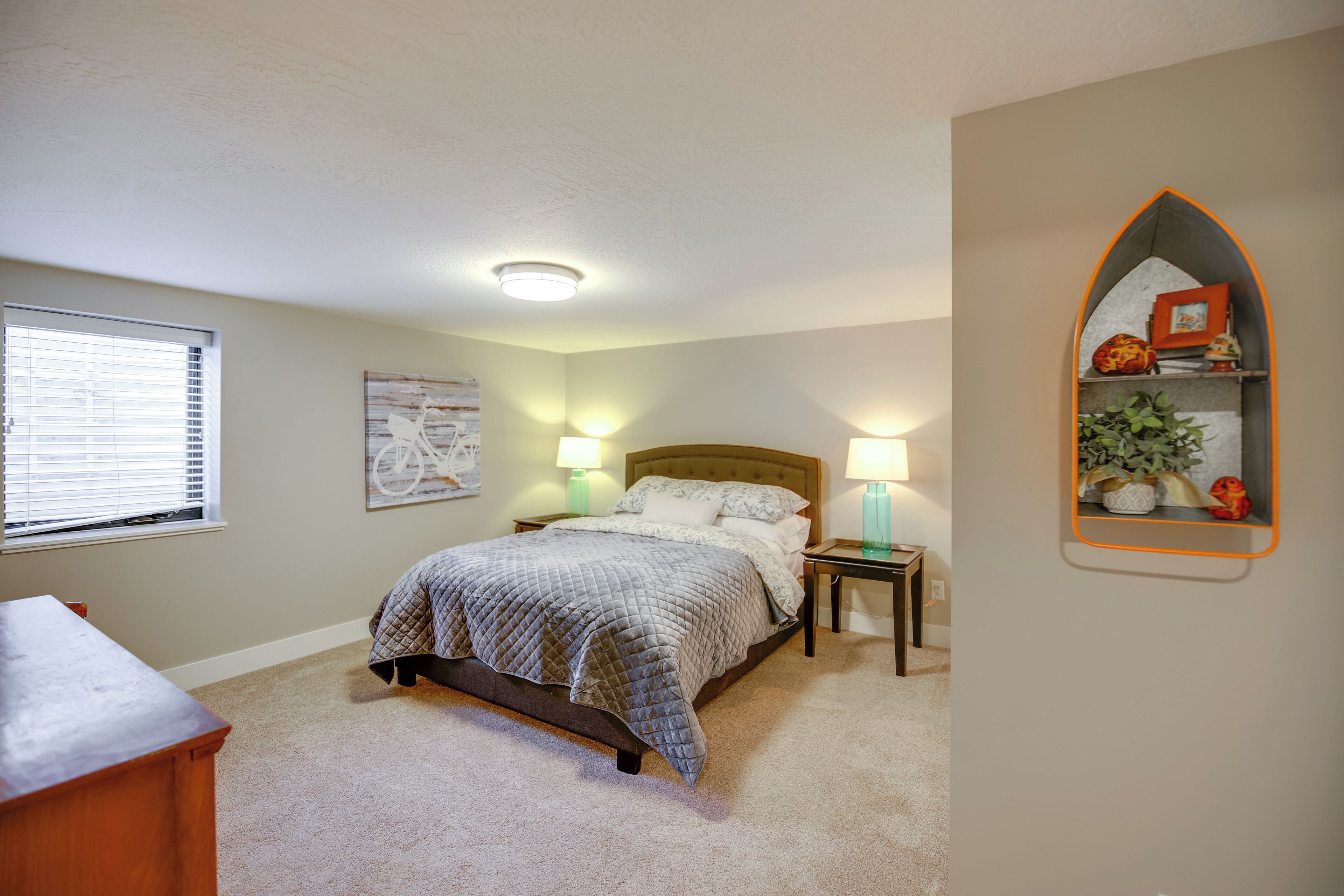 Une chambre à coucher comprenant un lit, des lampes de chevet, une table de chevet, une commode, une fenêtre avec des stores et une étagère murale décorative.