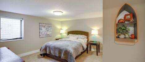 A bedroom with a bed, bedside lamps, a nightstand, a dresser, a window with blinds, and a wall-mounted shelf with decorative items.