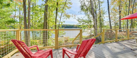 Eine Holzterrasse mit zwei roten Stühlen und einem roten Sonnenschirm mit Blick auf einen von Bäumen umgebenen See.