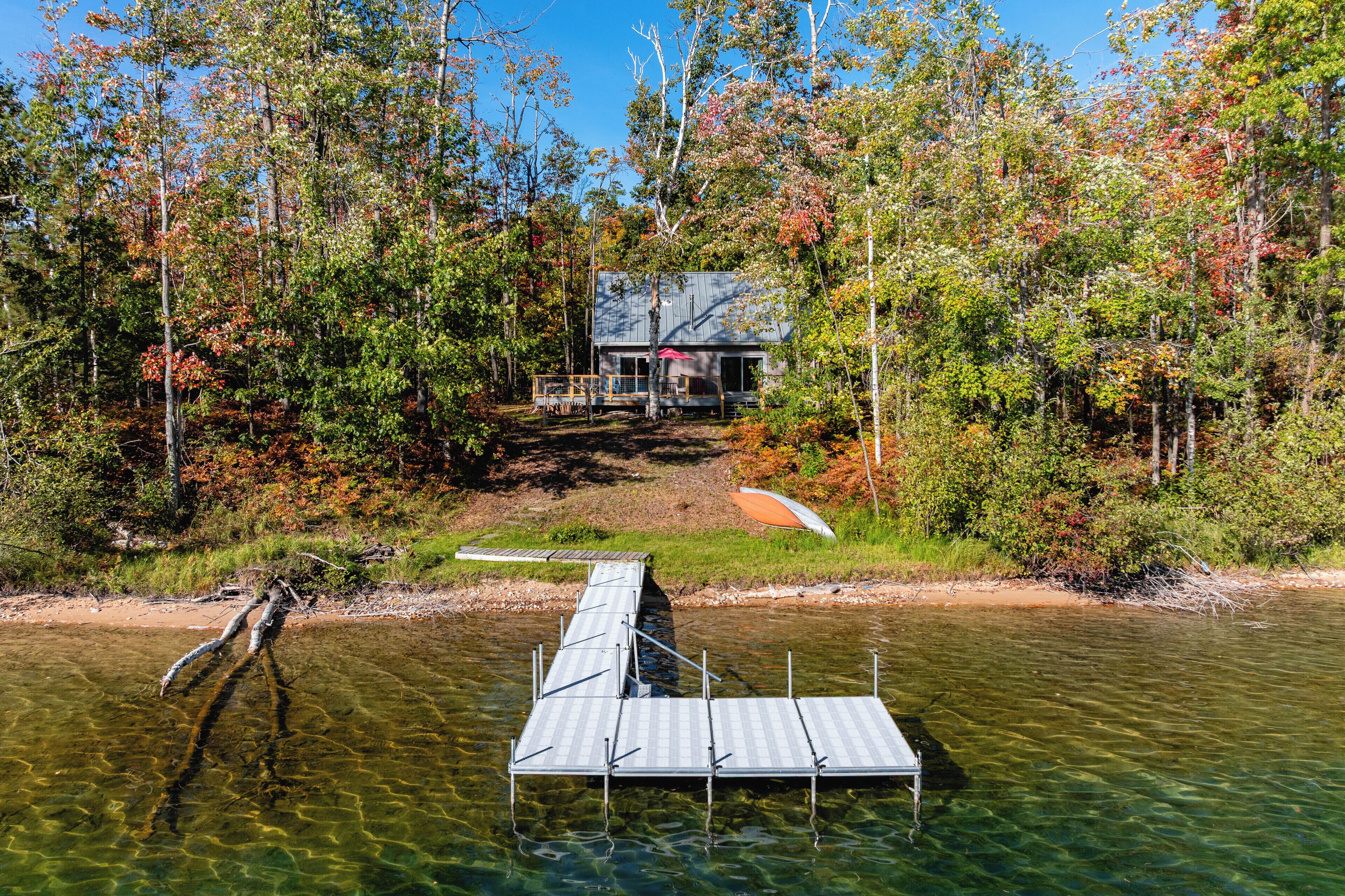 Une cabane au bord d’un lac aux eaux limpides, entourée d’arbres aux feuilles d’automne.