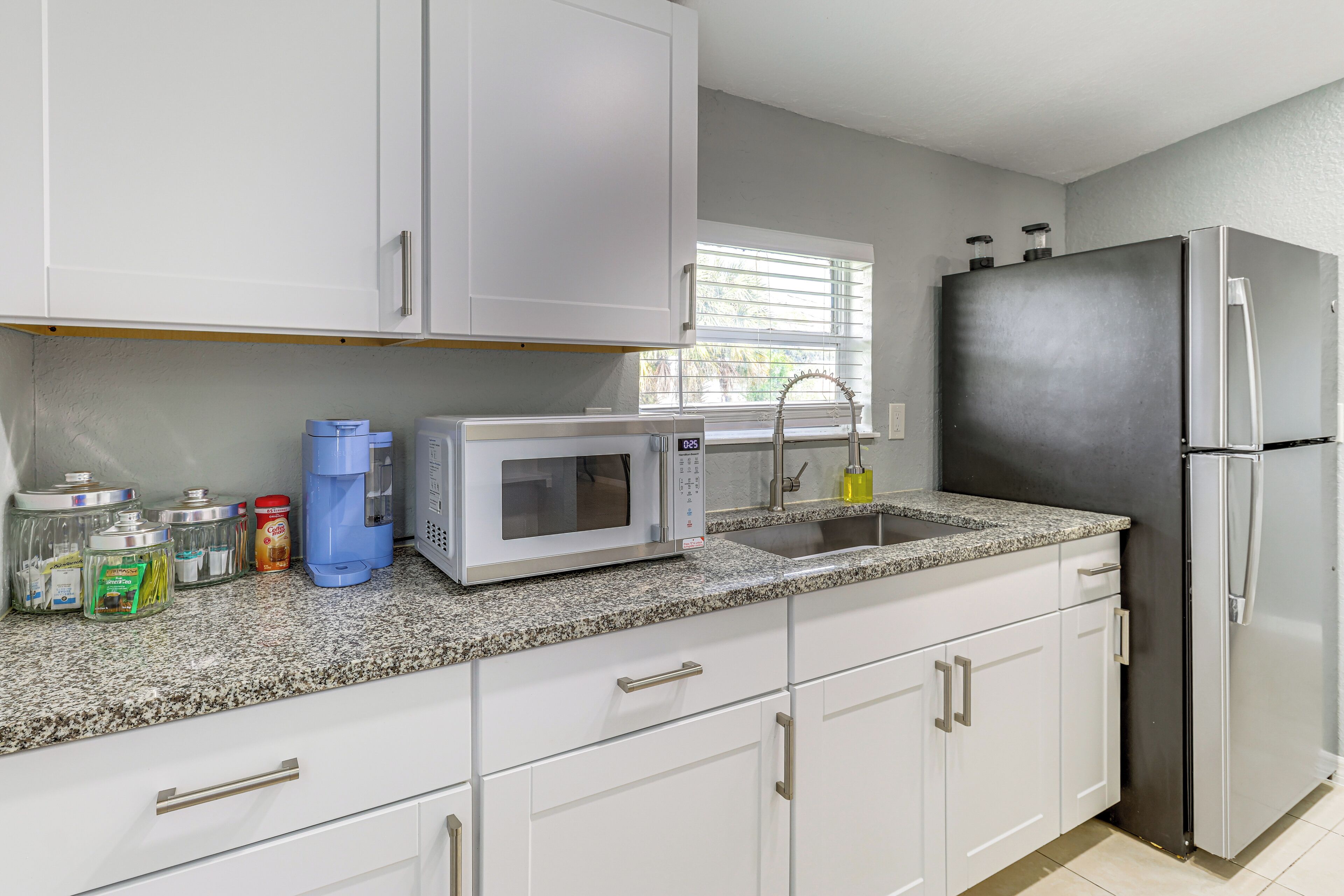 A kitchen with white cabinets, a granite countertop, a microwave, a refrigerator, and a window with blinds.