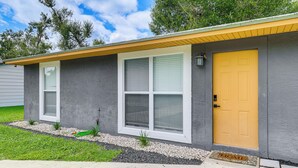Une maison de plain-pied avec une porte d’entrée jaune, de grandes fenêtres et un petit jardin à l’avant, agrémenté d’un chemin de gravier et de plantes.