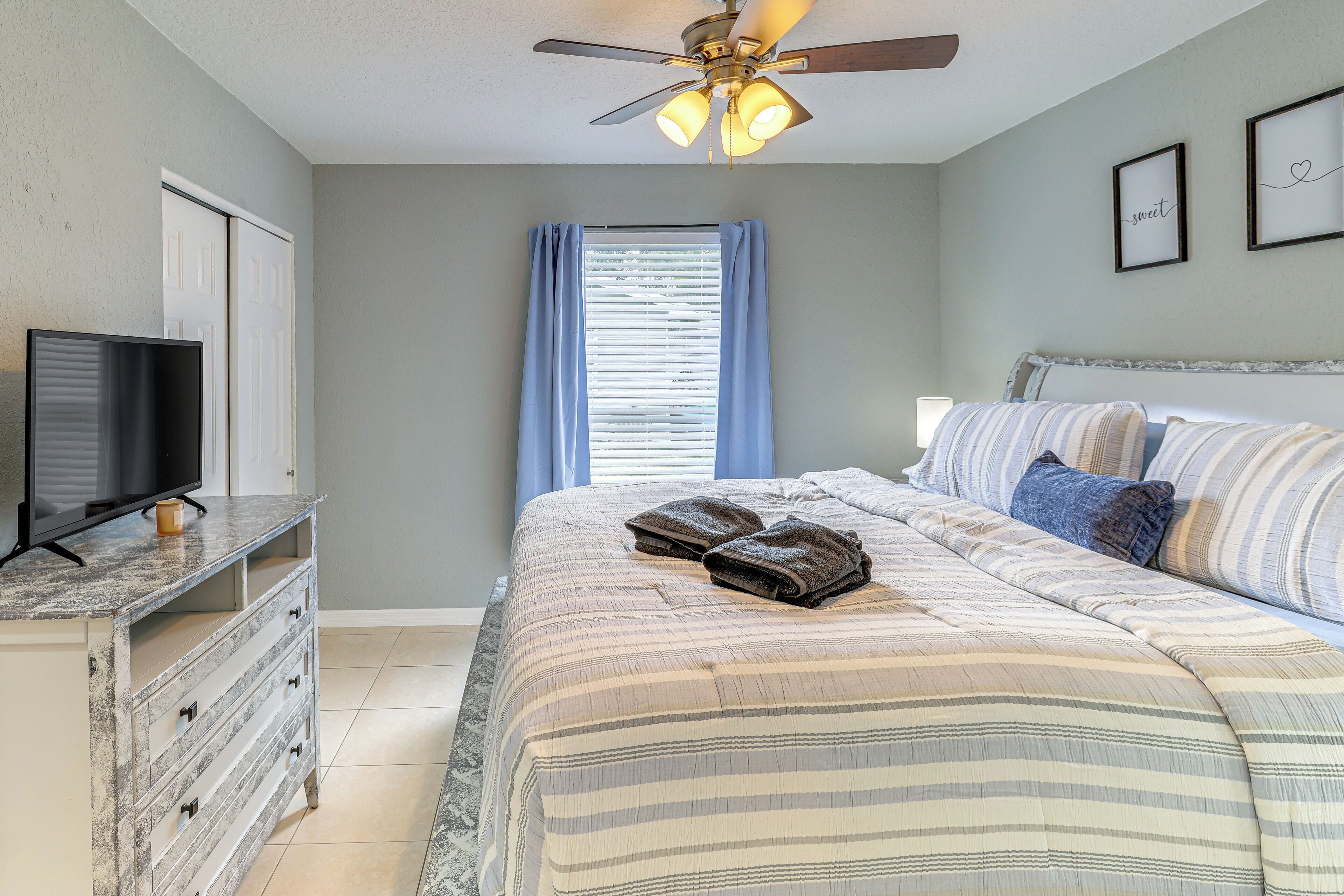 A bedroom with a bed, a TV on a dresser, a ceiling fan, and a window with curtains.