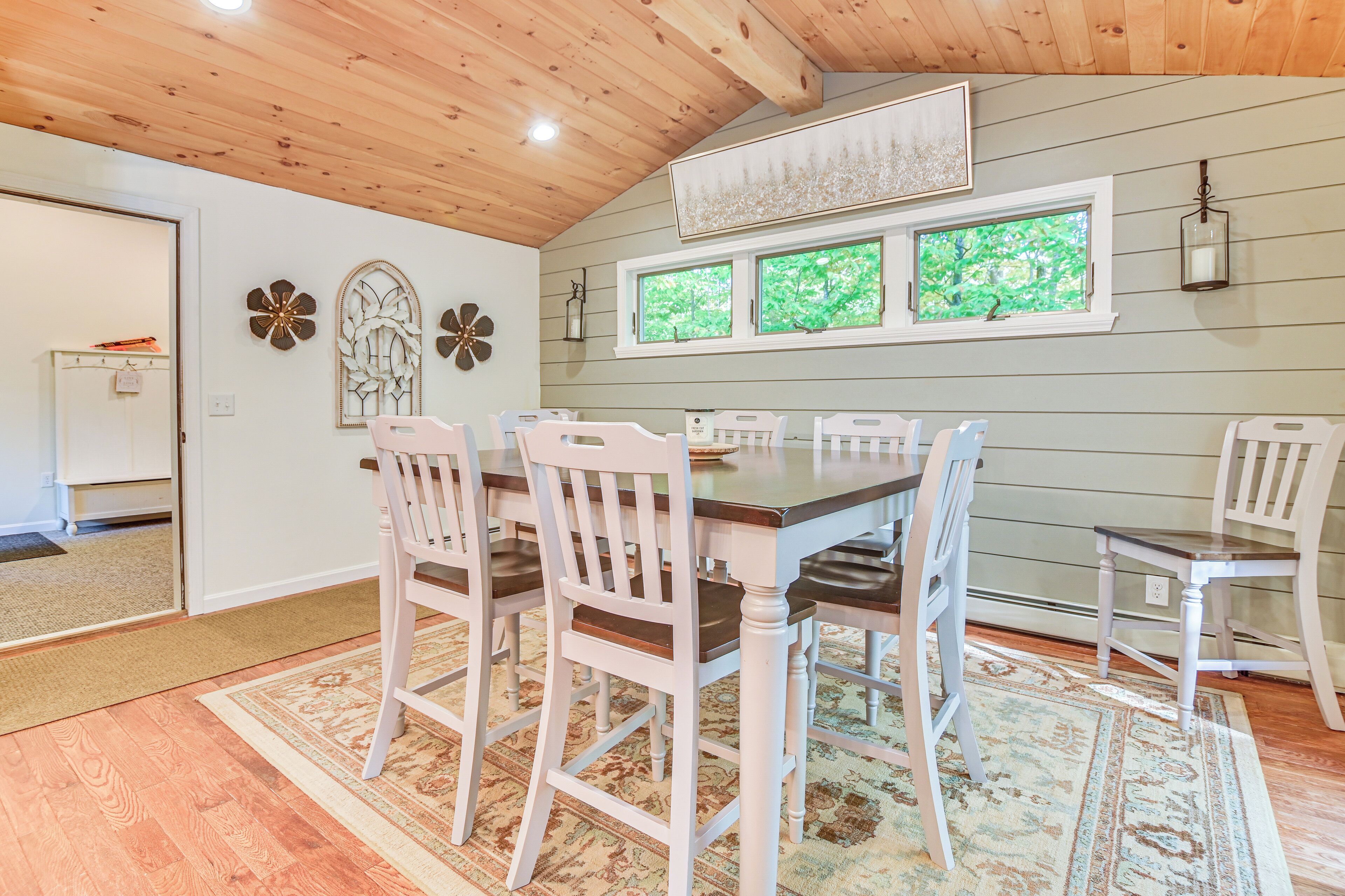 A dining area with a wooden table and white chairs, a window with a view of greenery, and a decorative wall piece.