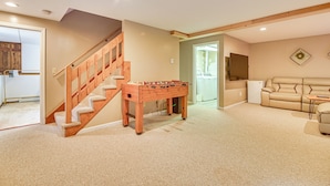 A spacious living area with a wooden staircase, a foosball table, a beige sofa, and a flat-screen TV mounted on the wall. - Hike, Leaf-peep, Ski: Hideaway w/ Private Hot Tub! (Intervale)