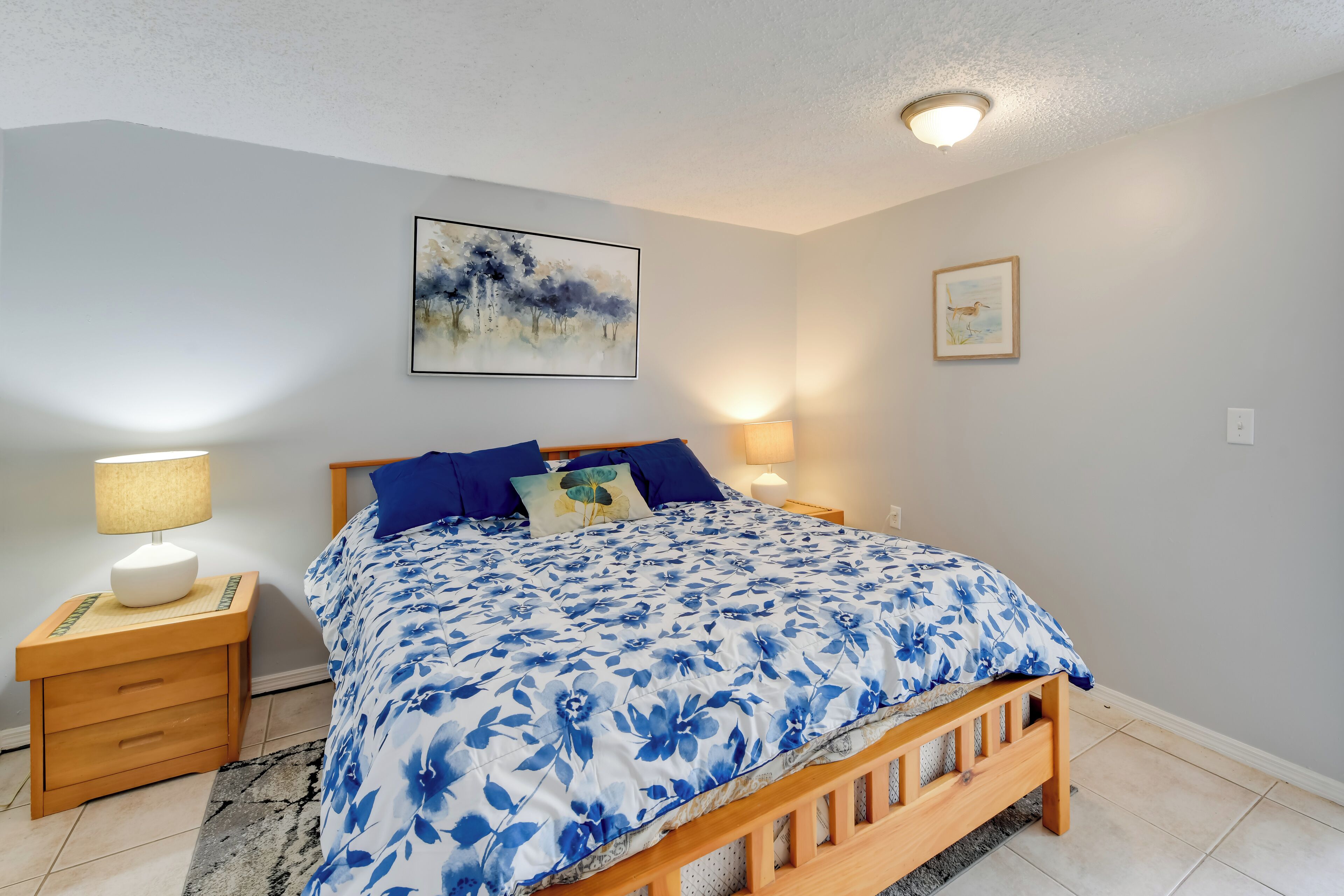 A bedroom with a wooden bed, blue floral bedding, two bedside lamps, a framed painting, and a rug.