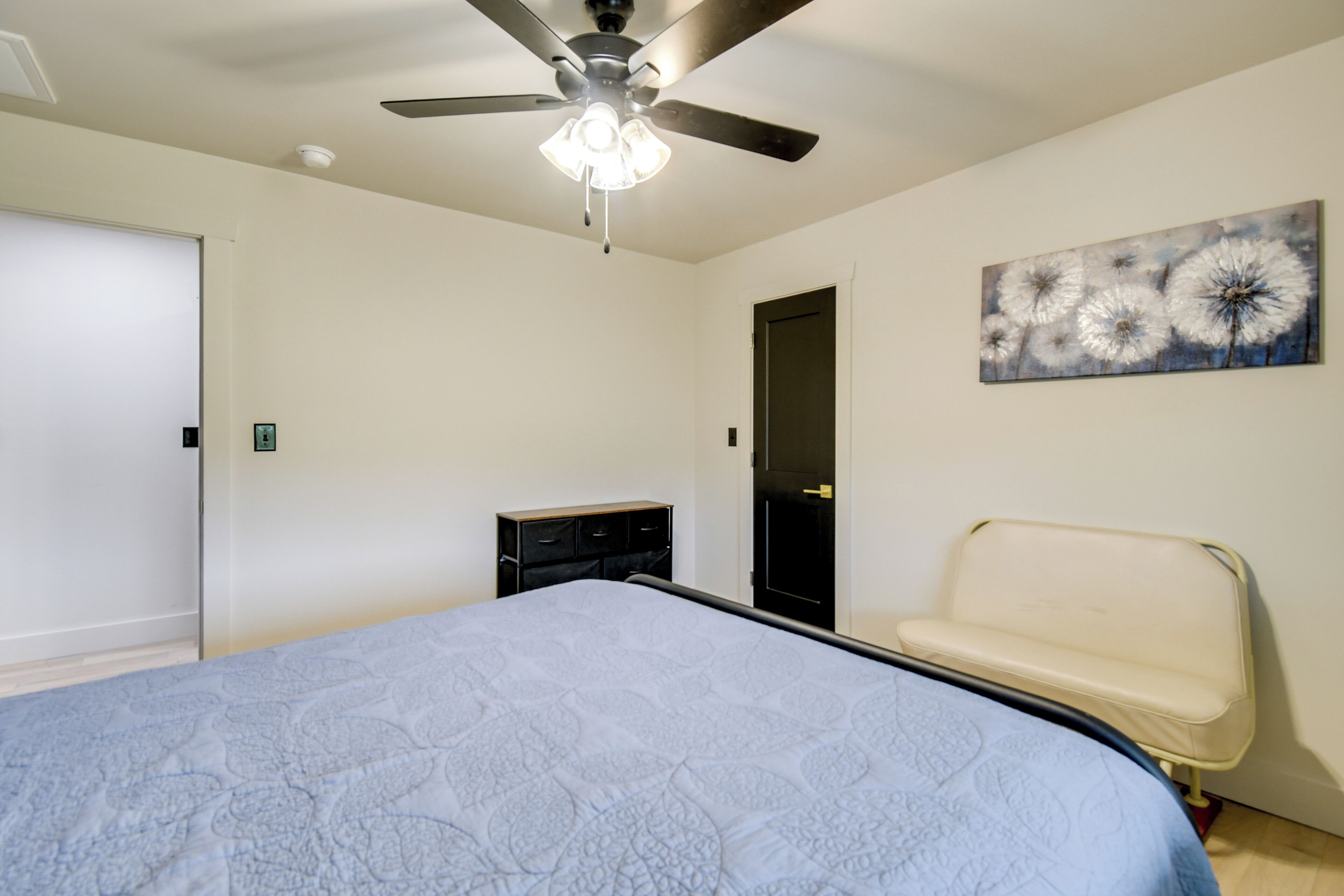 A bedroom with a bed, a ceiling fan, a dresser, a bench, and a dandelion painting on the wall.