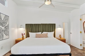 A hotel room with a bed, two bedside tables, a ceiling fan, a large abstract painting, and a mirror. - Deluxe King Studio in Boutique Hotel | The Rosalie (Palm Springs)