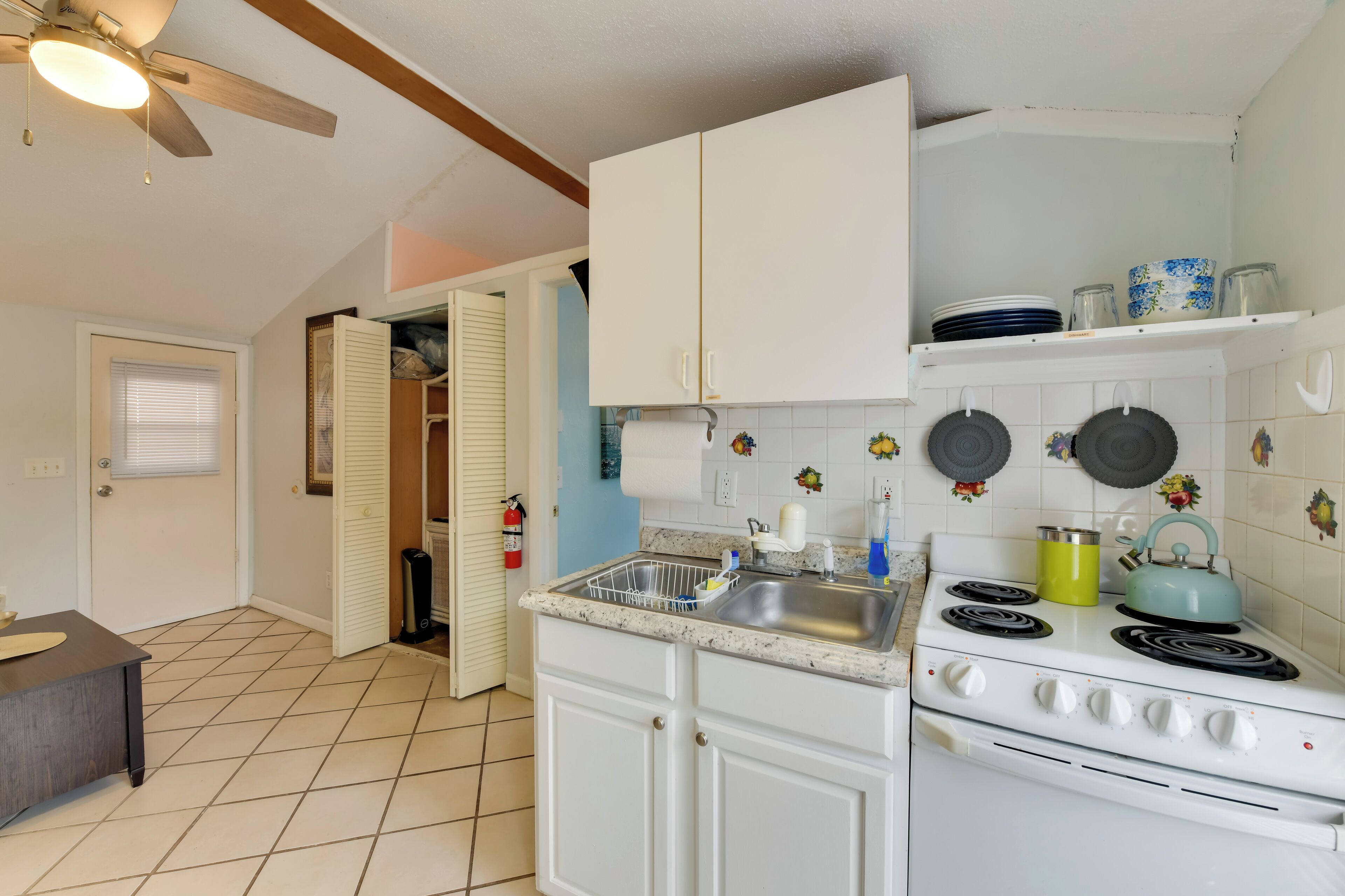 A kitchen with white cabinets, a stove, and a sink. There is a ceiling fan, a window with blinds, and a door leading to another room.