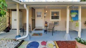 A covered porch with a welcome mat, two chairs, a potted plant, and a small table.
