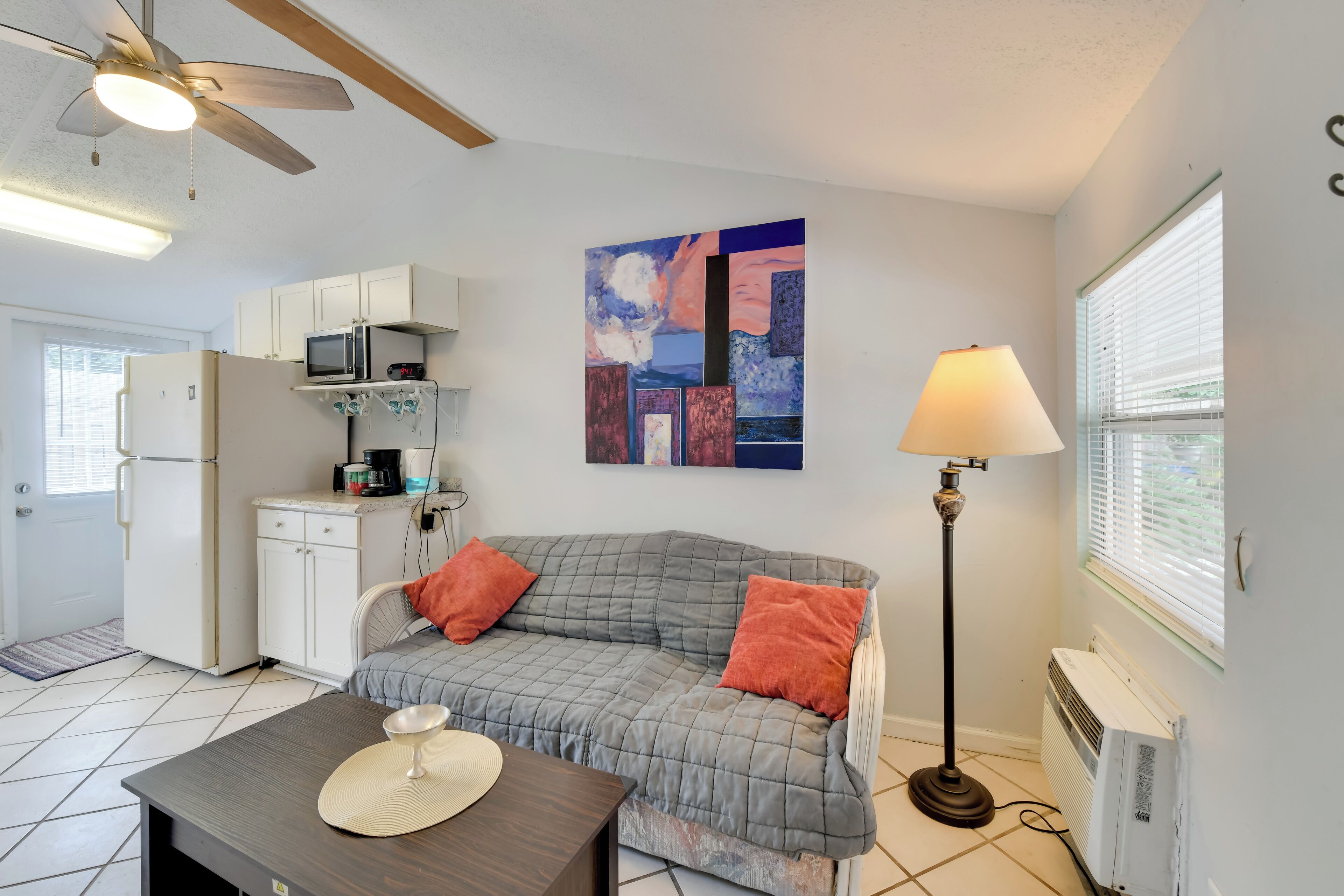A compact living space with a kitchenette, a gray couch with orange cushions, a dining table with a bowl and a mat, a standing lamp, and a ceiling fan.