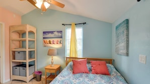 A bedroom with a bed, a nightstand, a lamp, a window with curtains, a ceiling fan, and a wall with a painting.