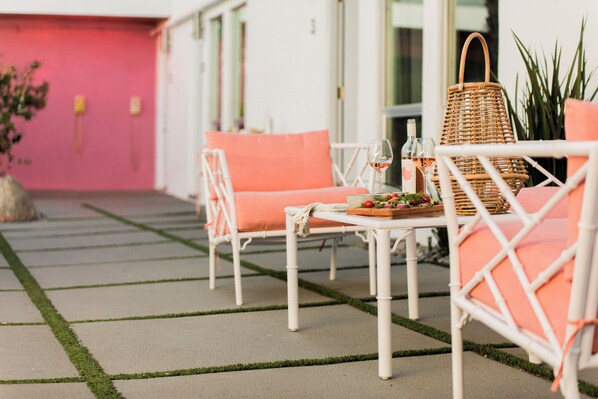 Un espace extérieur aménagé avec des meubles en osier blanc et des coussins orange, un panier en osier et une table sur laquelle se trouvent une bouteille et des verres.