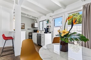 Une cuisine moderne avec des meubles blancs, un four à micro-ondes et une machine à café. On y trouve un coin repas avec une table ronde et des chaises, ainsi qu’une grande fenêtre avec des rideaux.