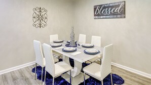 Comedor con una mesa blanca dispuesta para seis, sillas blancas, un adorno en la pared y una alfombra azul.
