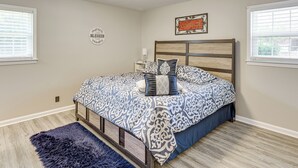 Un dormitorio con cama, alfombra, mesita de noche y un adorno en la pared.