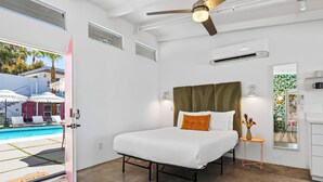 Ein Schlafzimmer mit Bett, Deckenventilator, Klimaanlage, einem kleinen Tisch mit einer Vase und Blick auf einen Swimmingpool.