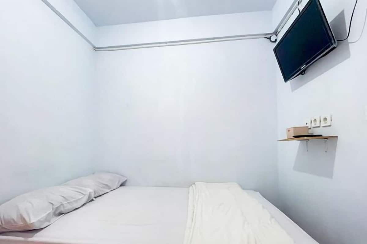 A small, white, single-bed room with a mounted TV and a shelf with a box.