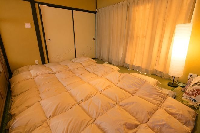 Quarto de hotel com uma cama grande, uma mesa de cabeceira com abajur e uma janela coberta por cortinas.