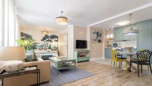 A modern living room with a sofa, coffee table, and a TV. There is a dining area with a table and chairs. The room has wooden flooring and light blue cabinets. - Cathedral Keepers - Exclusive apartment in Jerez (Jerez de la Frontera)