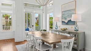 Een eethoek met een houten tafel, witte stoelen, een kroonluchter en een schilderij van een zeegezicht.