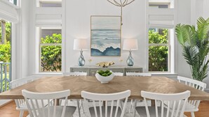Een eethoek met een houten tafel, witte stoelen, een decoratieve middelpunt met groene planten en een schilderij van de oceaan aan de muur.