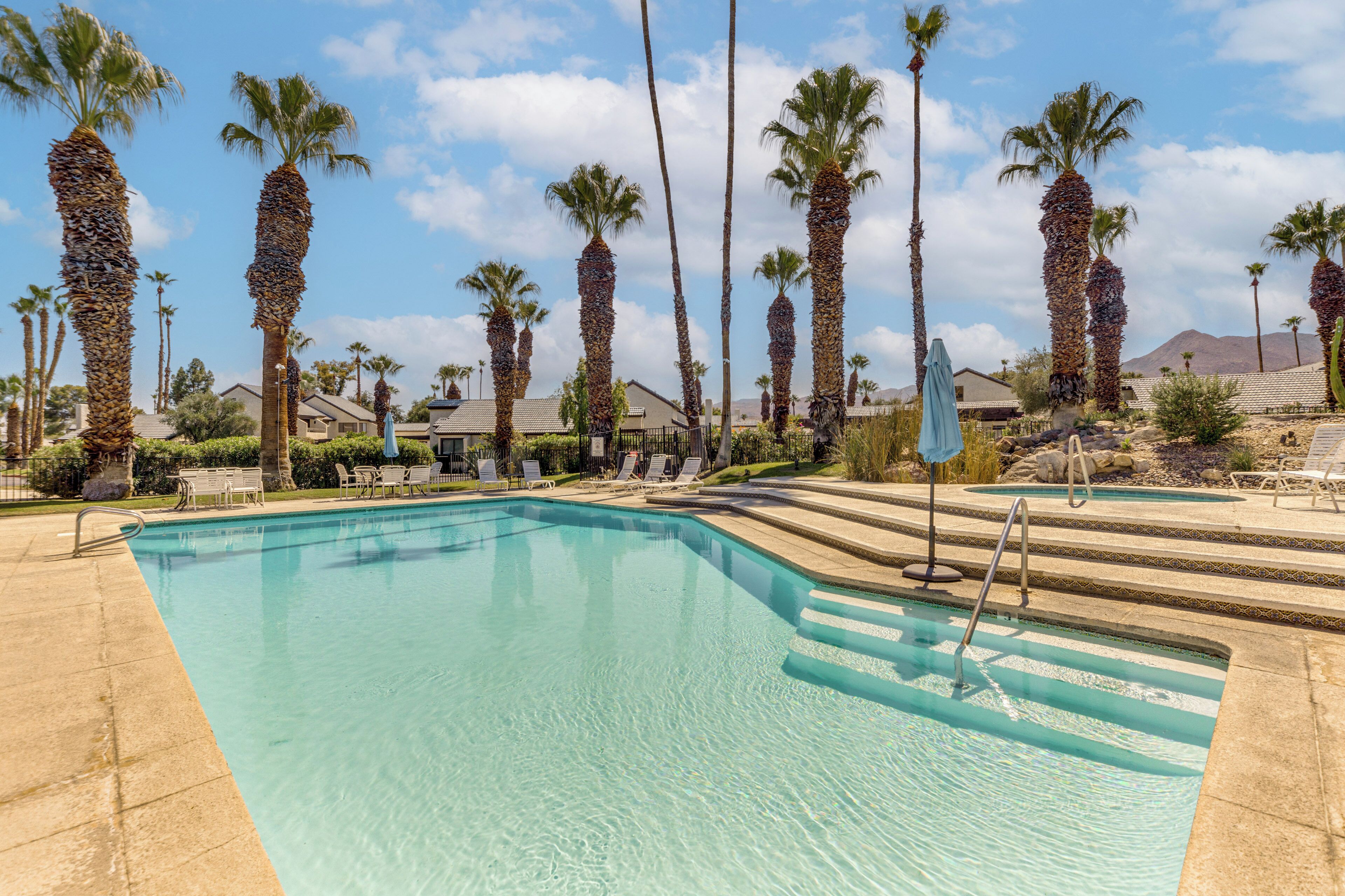 A clear swimming pool with steps leading into it, surrounded by palm trees and lounge chairs.