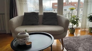A living room with a beige sofa, two dark cushions, a small round table with a decorative item, and a view of buildings through large windows. - Meszko Park House Budapest (Budapest)