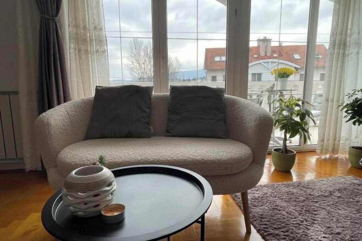 Een woonkamer met een beige bank, twee donkere kussens, een klein rond tafeltje met een decoratief object, en een uitzicht op gebouwen door grote ramen.