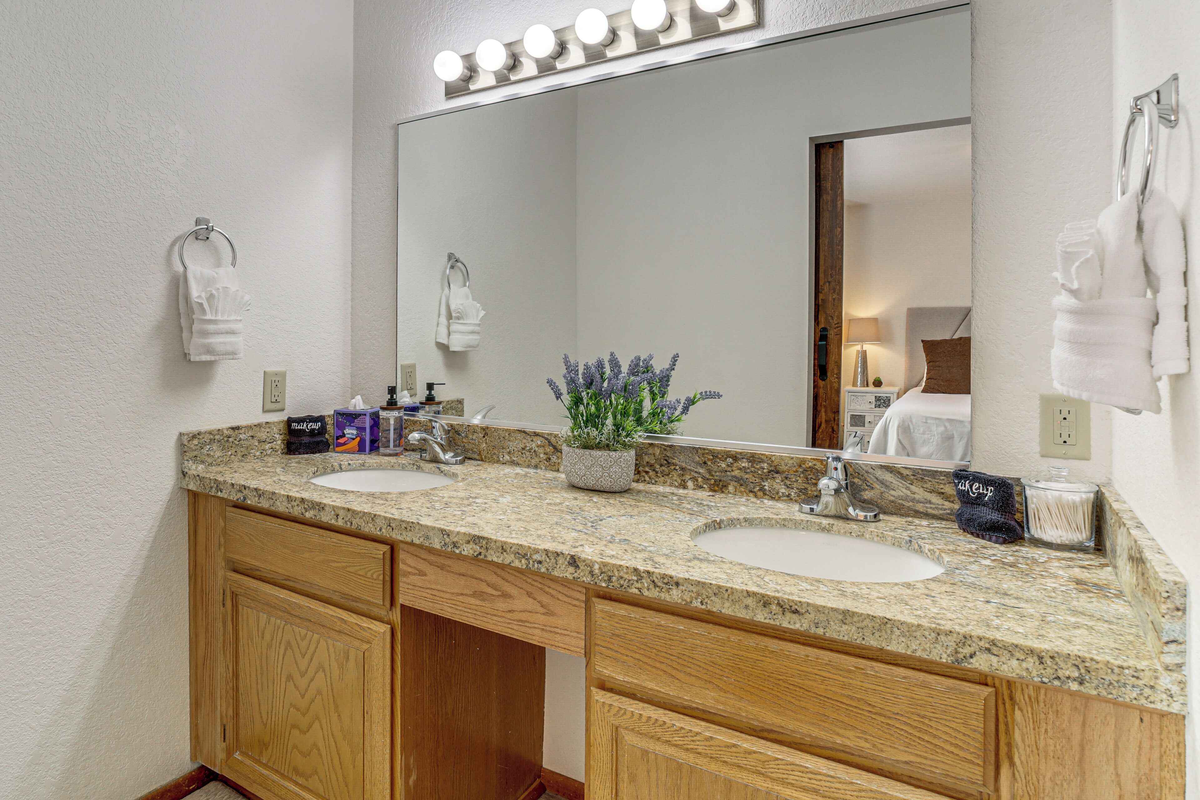 A bathroom with two sinks, wooden cabinets, a large mirror with lights, and a potted plant.