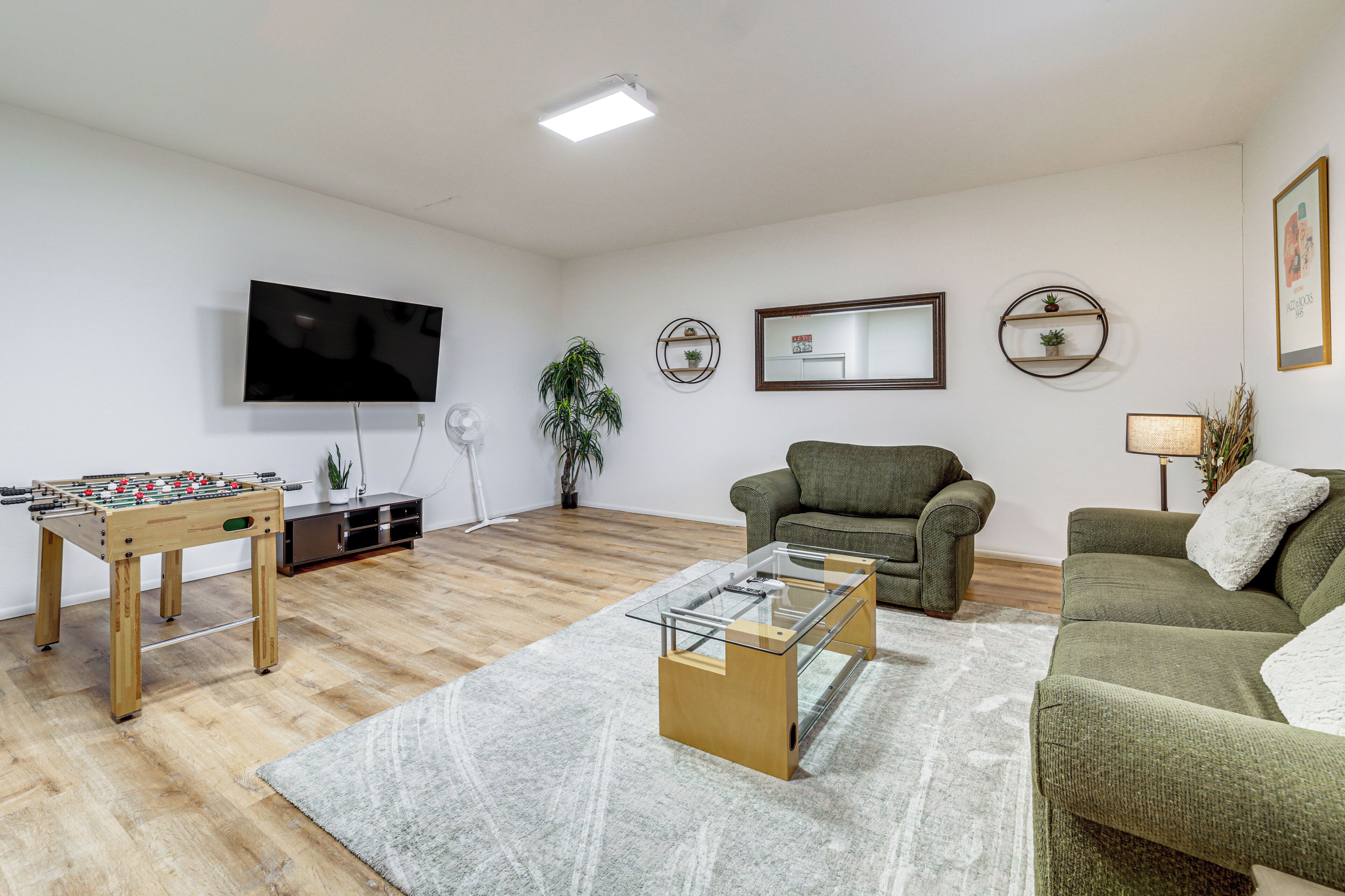 Una sala de estar con una mesa de fútbol de mesa, una mesa de centro de vidrio, un sofá, un televisor y objetos decorativos en las paredes.