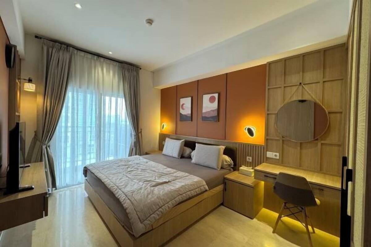 A modern hotel room with a large bed, bedside tables, a desk with a chair, and artwork on the walls.
