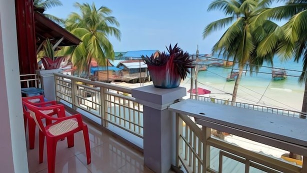 A balcony with red plastic chairs, a large potted plant, and a view of a beach with palm trees and boats. - Island Boutique Koh Rong (Sihanoukville)
