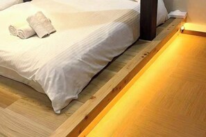A neatly made bed with a white comforter and rolled towels, a wooden floor, and a wall-mounted light fixture. - JIA MING (Osaka)