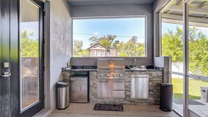 An outdoor kitchen with a grill, oven, and sink, a trash can, and a view of a house through a large window.