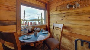 L’intérieur d’un chalet en bois, avec une table à manger dressée pour deux personnes, une fenêtre donnant sur une zone boisée et un luminaire fixé au mur.