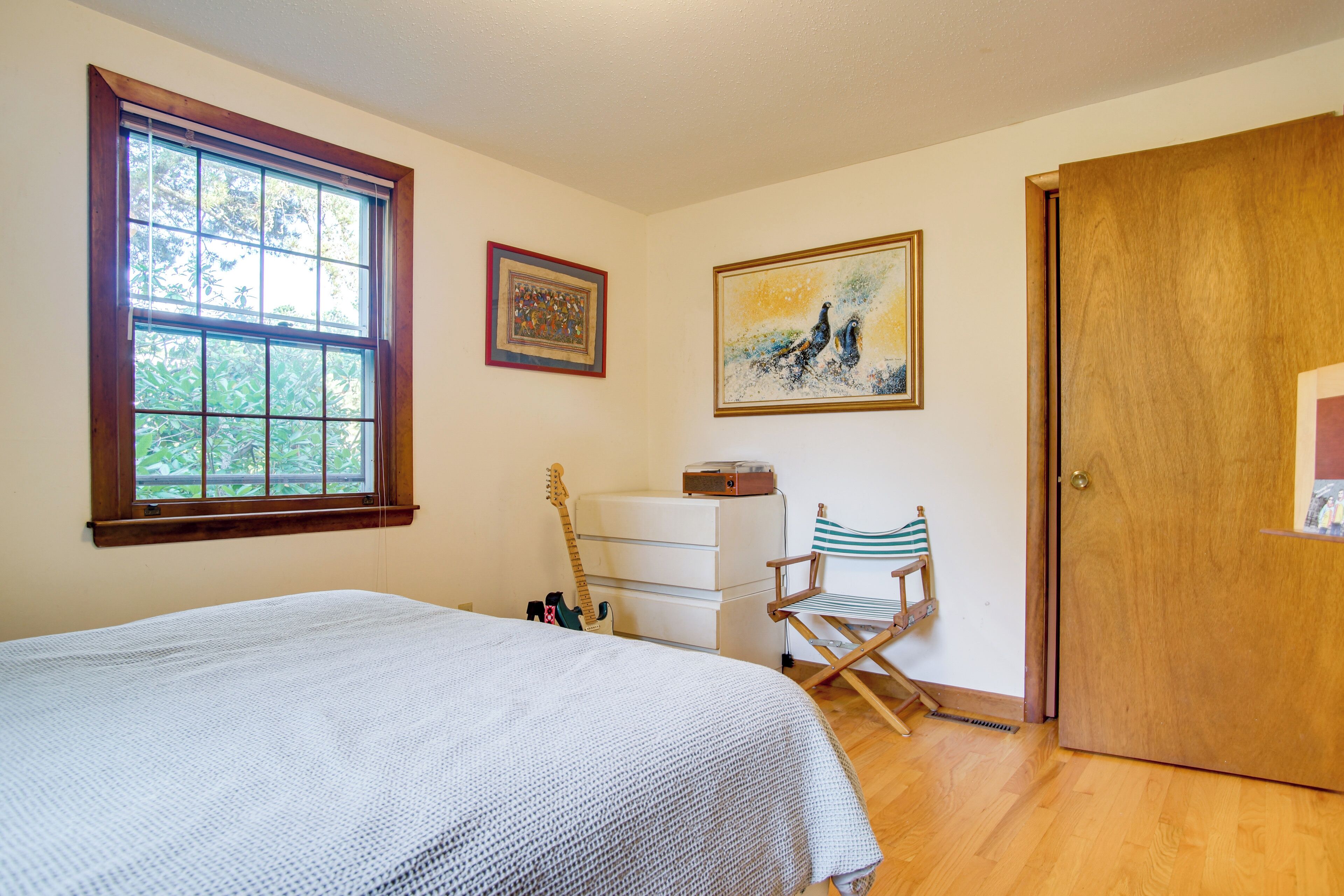 Ein Schlafzimmer mit großem Fenster, einem Bett, einem Schminktisch, einem Sessel und zwei gerahmten Kunstwerken an der Wand.