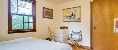 Ein Schlafzimmer mit großem Fenster, einem Bett, einem Schminktisch, einem Sessel und zwei gerahmten Kunstwerken an der Wand.