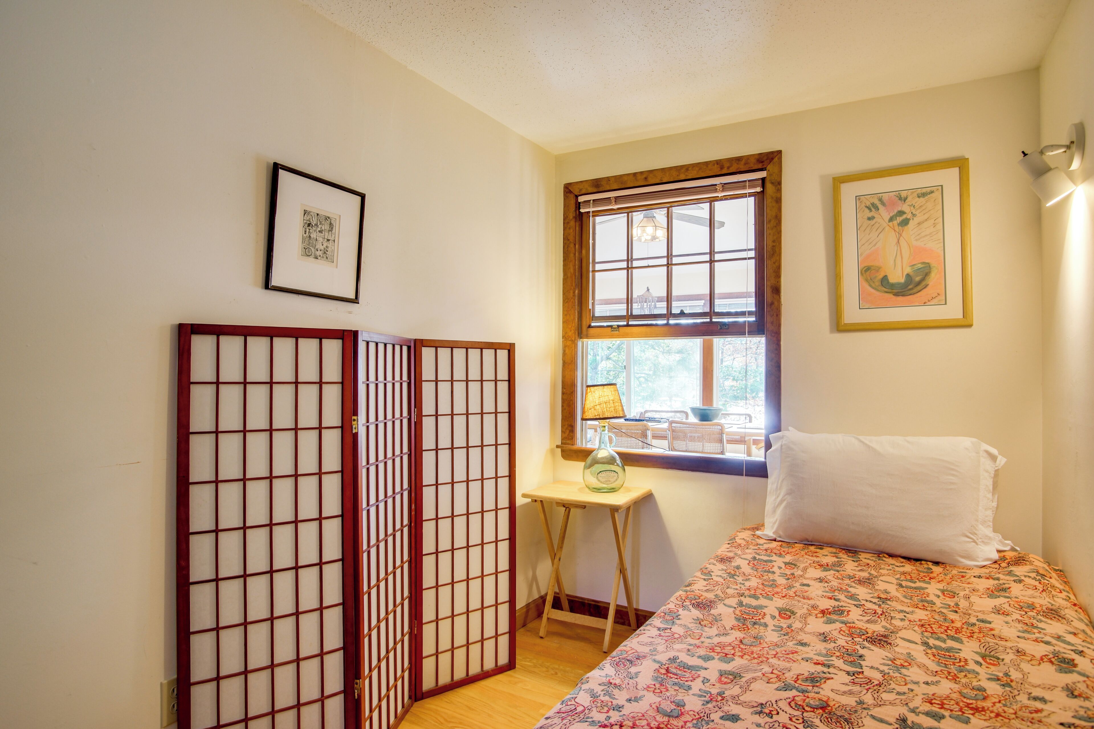 Una habitación con una cama, una mesita con lámpara, una ventana con persianas y un cuadro en la pared.