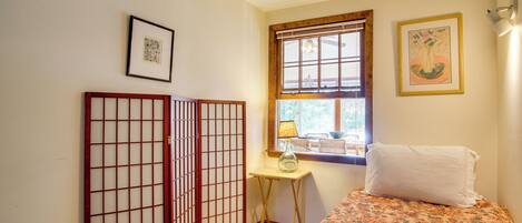 Una habitación con una cama, una mesita con lámpara, una ventana con persianas y un cuadro en la pared.
