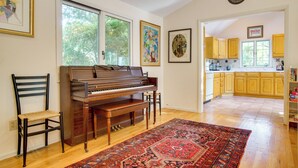 A piano in a room with a large window, a rug, and a kitchen in the background.