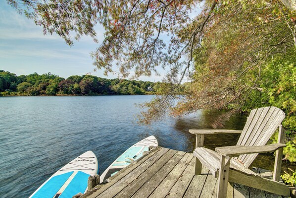 A wooden dock with a paddleboard and a chair by a calm lake. - 4,200 Sq Ft Mid-cape Cottage w/ Furnished Deck! (Brewster)