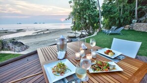 An outdoor dining table with plates of food, glasses of wine, and candles, overlooking a beach. - The View (Koh Samui)
