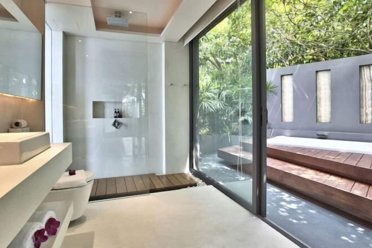 A modern bathroom with a glass shower, a wooden bench outside, and a view of greenery.