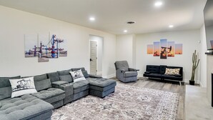 A living room with a sectional sofa, a grey armchair, a black sofa, and a patterned rug. There are wall art pieces depicting industrial and sunset scenes. - Backyard & Game Room! Home in Heart of Midland (Midland)