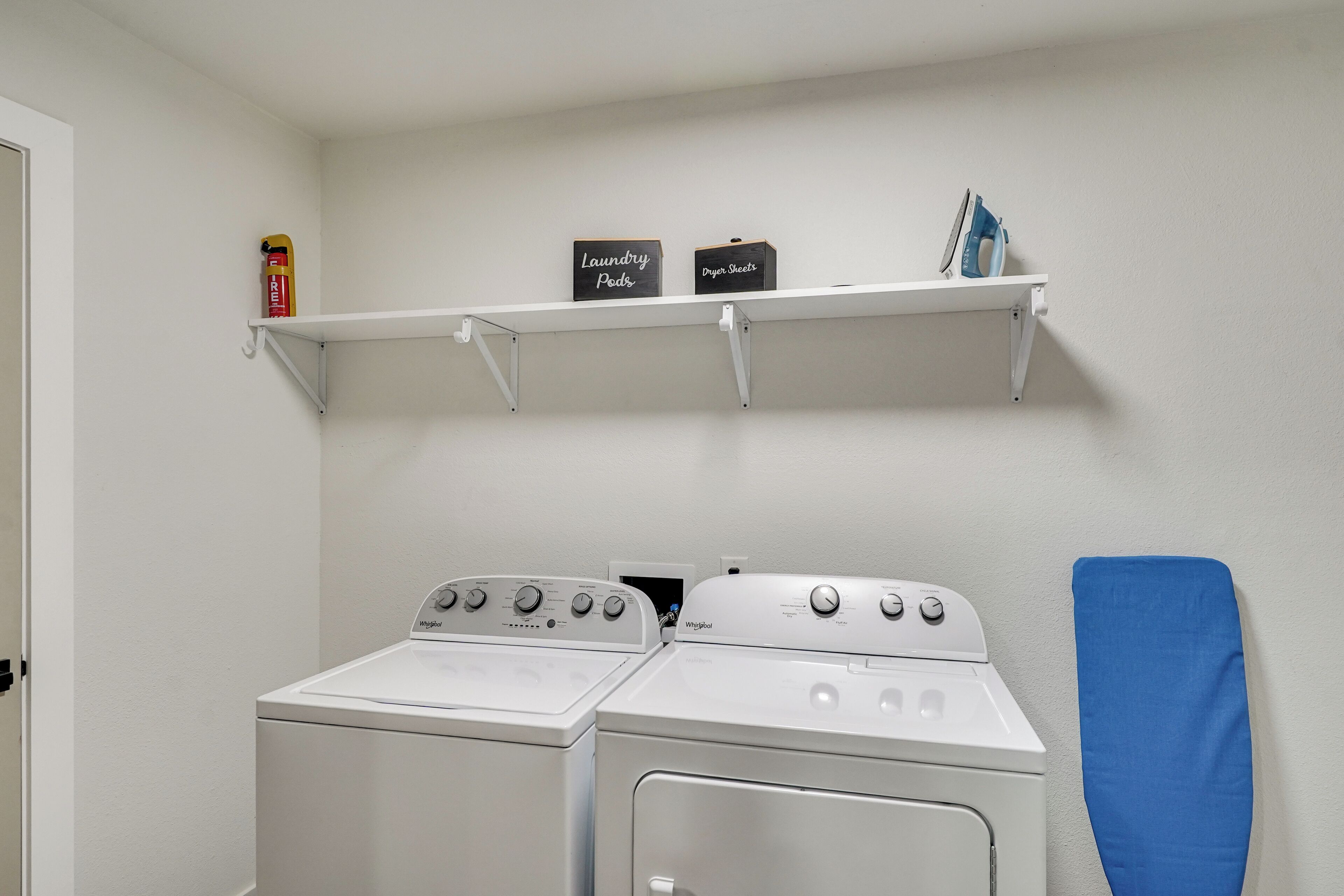 A laundry area with two Whirlpool dryers and a shelf above containing a fire extinguisher, laundry pods, and a box of dryer sheets.