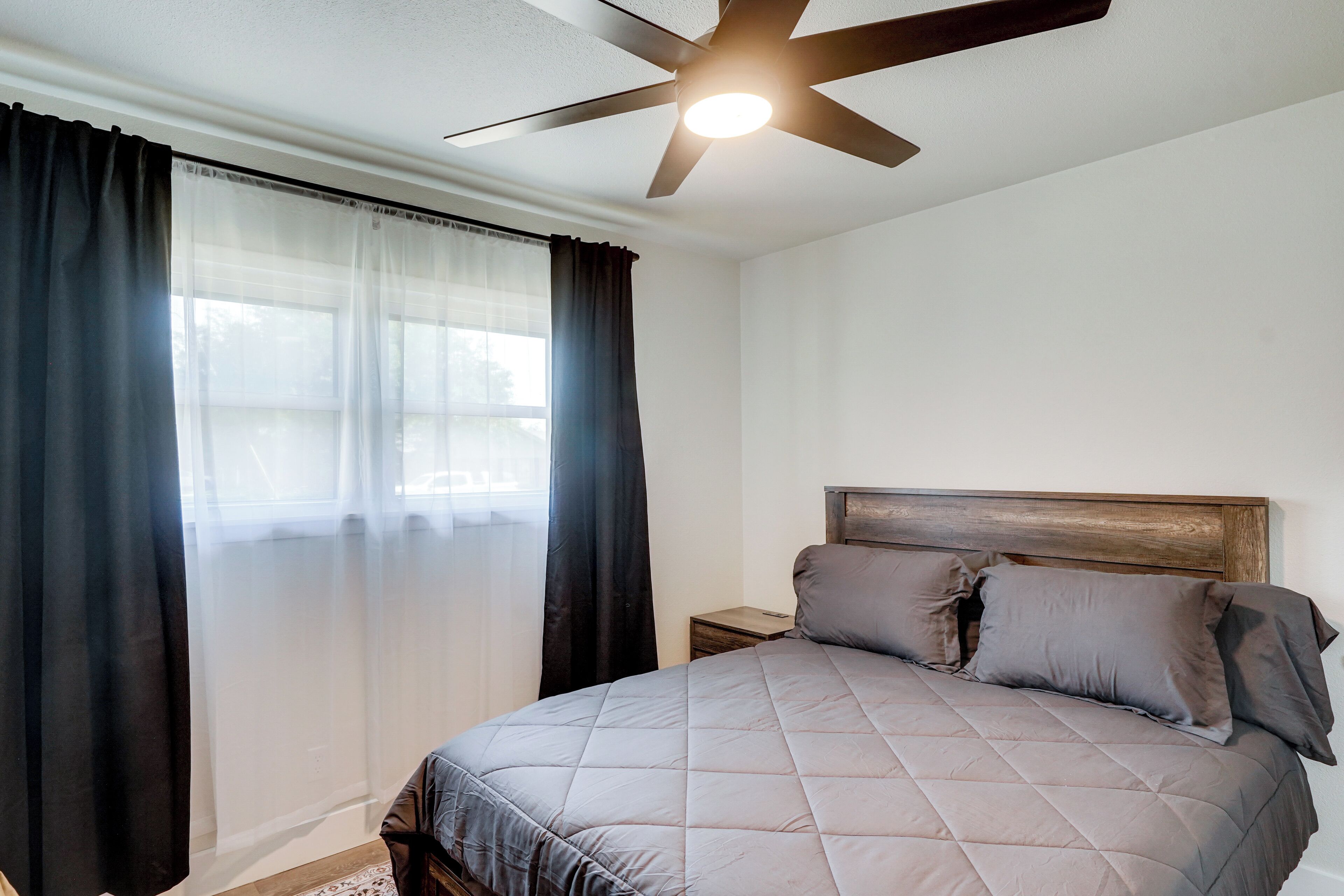 A bedroom with a bed, a nightstand, a ceiling fan, and sheer curtains.