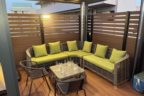 Un espacio exterior techado con mobiliario de mimbre, incluyendo un sofá y sillas, y una mesa de vidrio sobre piso de madera.