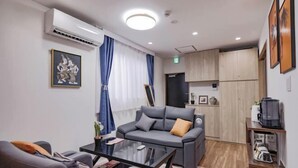 A modern hotel room with a sofa, a glass coffee table, and a kitchen area with wooden cabinets.