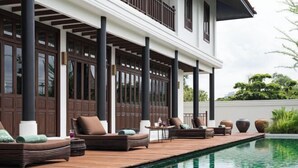 Un espace aménagé au bord de la piscine, avec des chaises longues, une terrasse en bois et un bâtiment doté de grandes fenêtres et de colonnes.